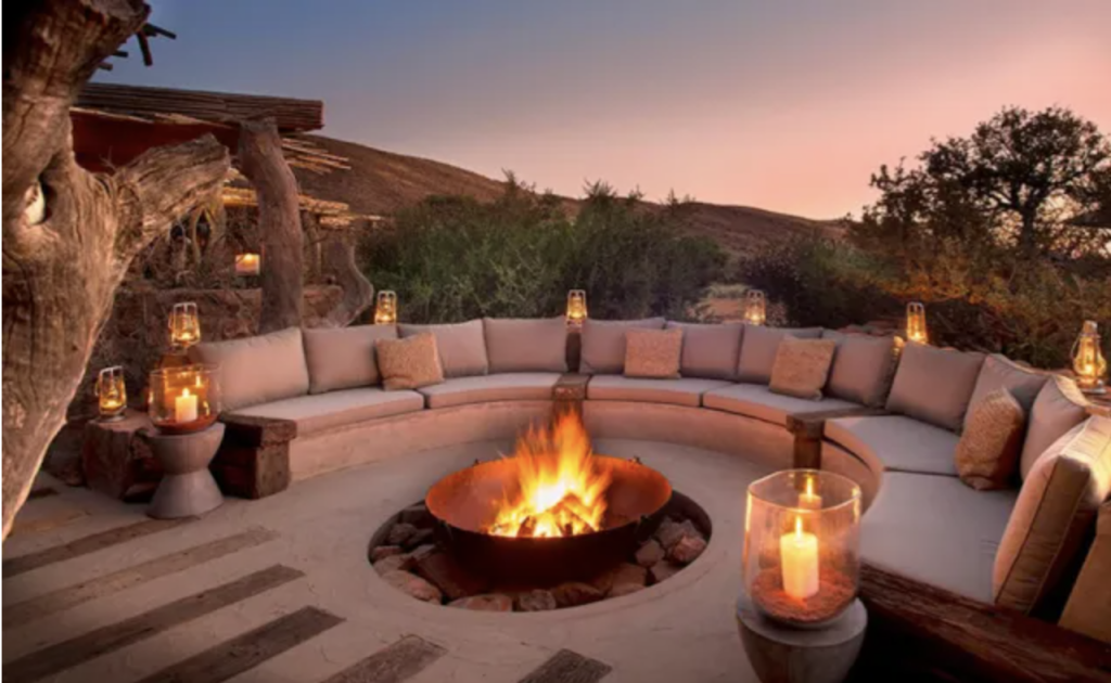 Outdoor lounge area around a fire fit with atmospheric lighting.