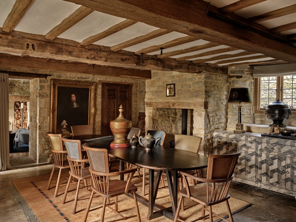 Dining room in a Cotswold stone farmhouse with exposed oak beams, an oval dark wood dining table, Windsor-style chairs and an inglenook fireplace